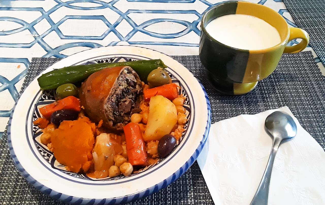 Ein Teller Couscous mit Fleisch-Osban, neben dem Teller liegt eine Serviette sowie ein Löffel und eine Tasse. Der Tisch ist mit einem blau-weißen geometrischen Linienmuster verziert.