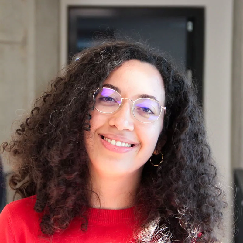 Portraitaufnahme unserer aus Sfax in Tunesien stammenden Projektmanagerin Mariem. Sie lächelt in die Kamera, trägt ein roten Sweater und eine Brille mit runden Gläsern.