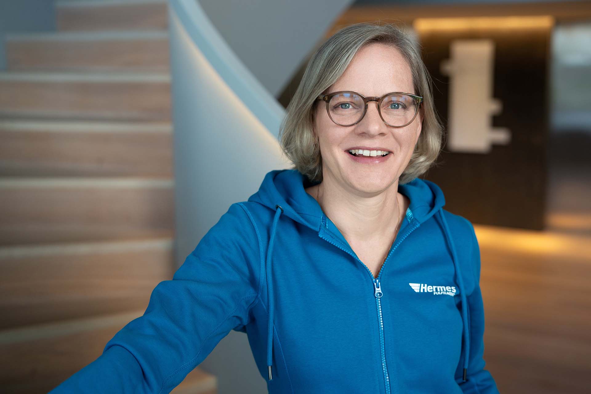 Barbara Schr&ouml;der&mdash;Senior Project Manager at Hermes Fulfilment. She is standing in front of a staircase in an office building, smiling directly at the camera. She is wearing a blue hoodie with the brand name &ldquo;Hermes Fulfilment&rdquo; printed in white letters in the upper right corner.