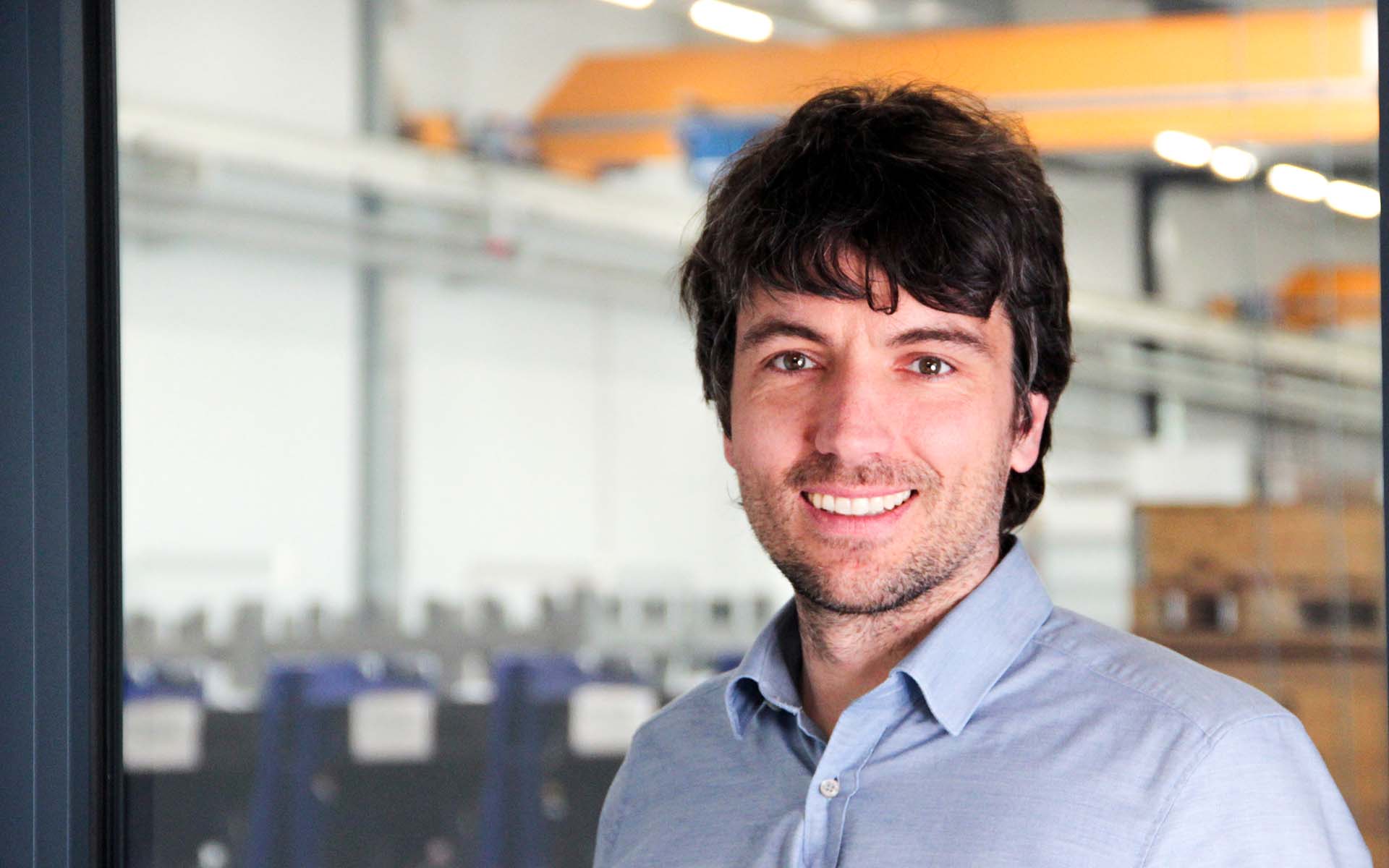Klaus J&auml;ger&mdash;Head of Special Projects and Modernization&mdash;smiles at the camera; in the background is a factory floor with a yellow overhead crane.