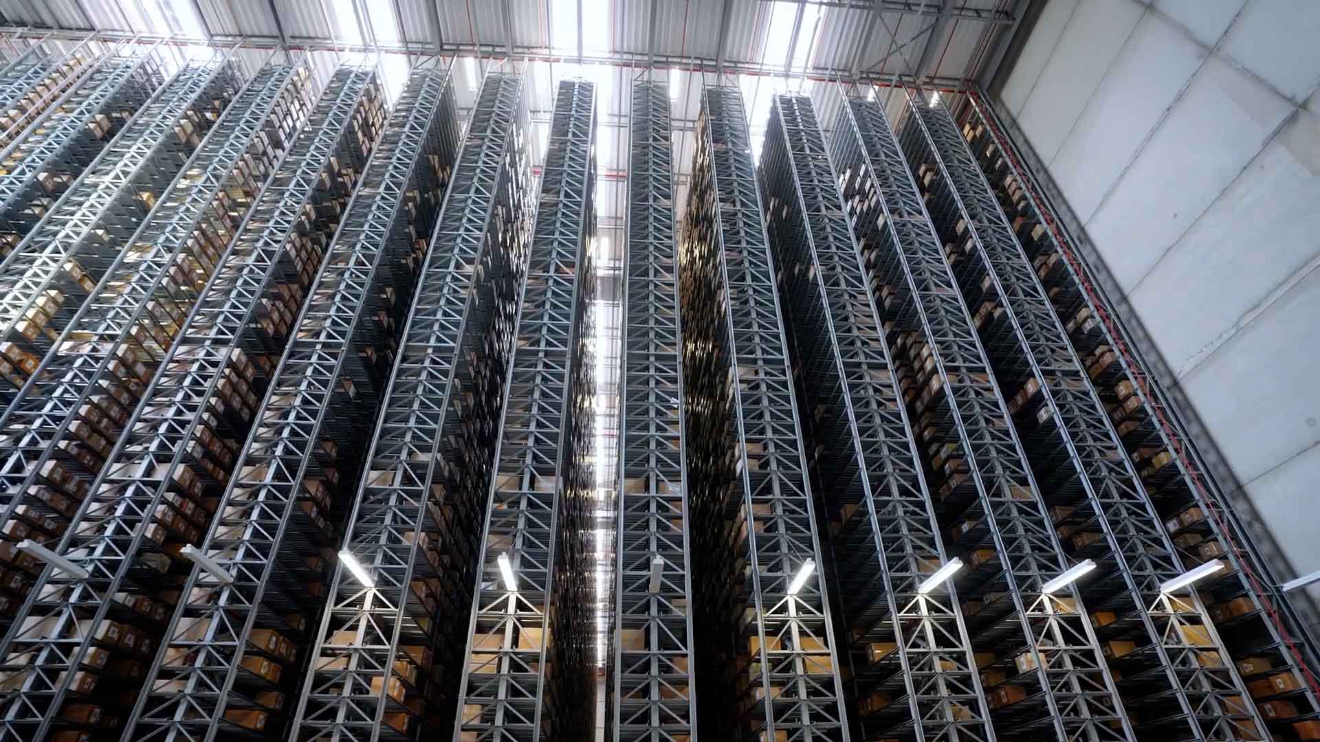 A ground-level view of the high-bay warehouse at the Hermes fulfillment center in Haldensleben. Several numbered high-bay racks and the warehouse ceiling are visible. The racks contain numerous boxes. Daylight streams into the warehouse through the skylights.