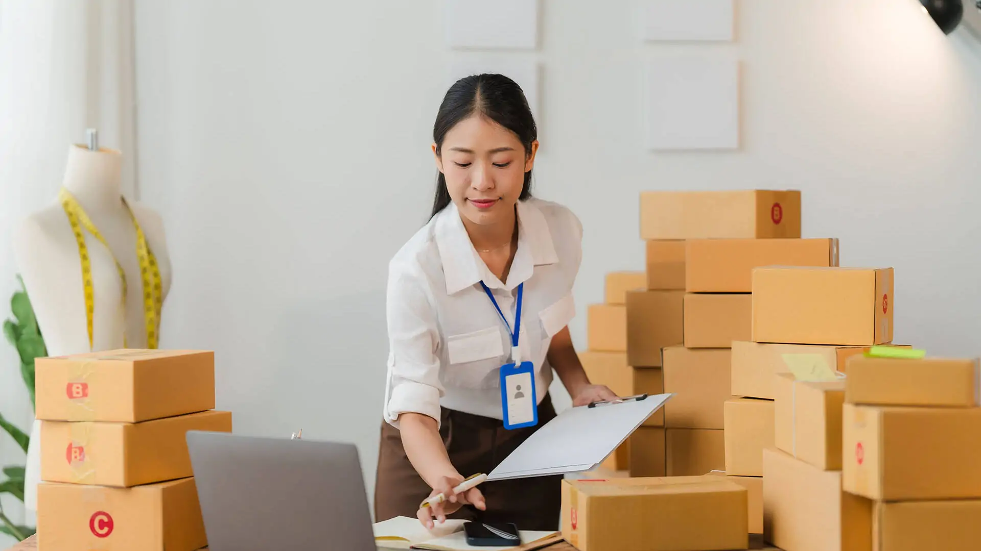 Asian SME entrepreneur meticulously manages online orders in her store, surrounded by shipping boxes