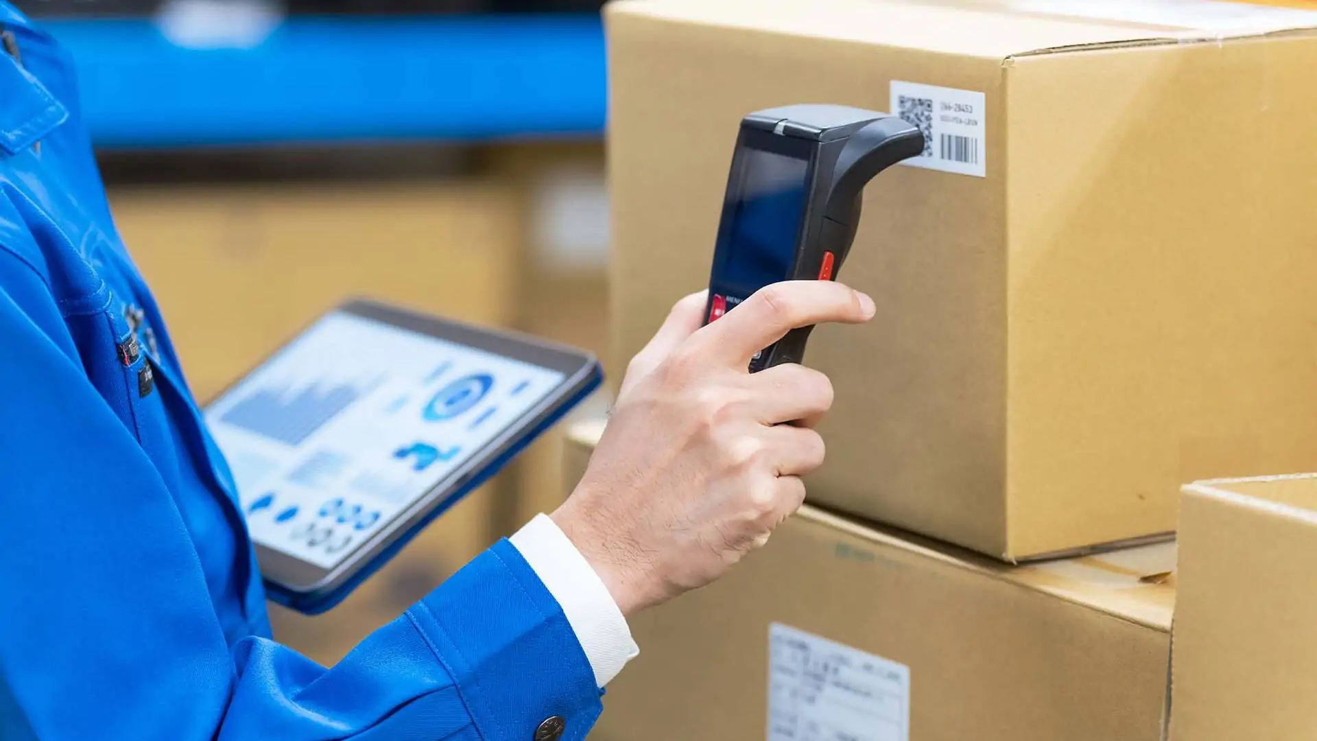 Mitarbeiter in blauer Arbeitskleidung, der ein mobiles Datenterminal hält, während er einen Barcode von einer Kiste in einem Lagerhaus scannt