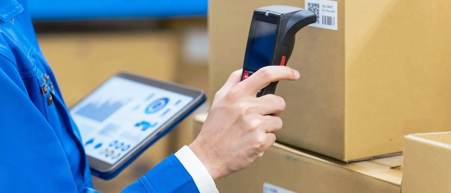 Mitarbeiter in blauer Arbeitskleidung, der ein mobiles Datenterminal hält, während er einen Barcode von einer Kiste in einem Lagerhaus scannt