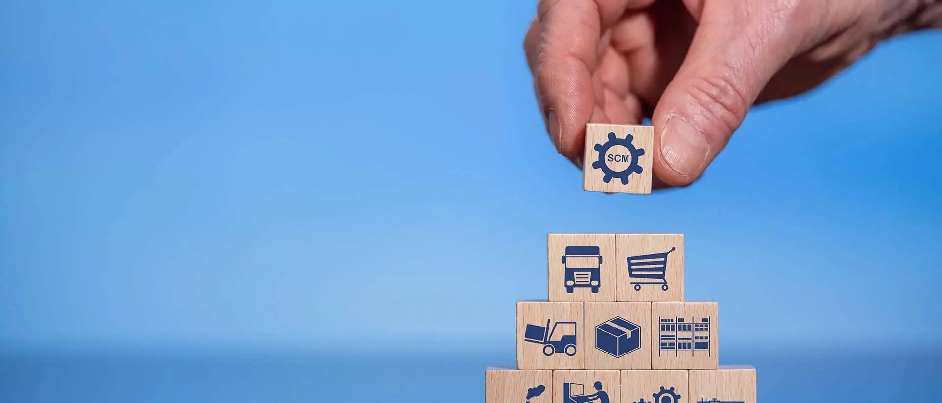 Hand, die einen Holzklotz mit einem Zahnradsymbol auf eine Pyramide aus Klötzen legt, die Symbole der Lieferkette darstellen, vor einem blauen Hintergrund.