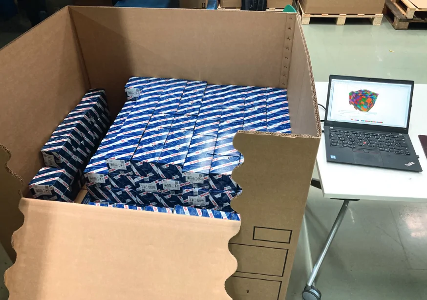 Blick in Karton mit Warenkartons, daneben Laptop auf Tisch, auf dem die Befüllung des Kartons in 3d vorsimuliert wird.