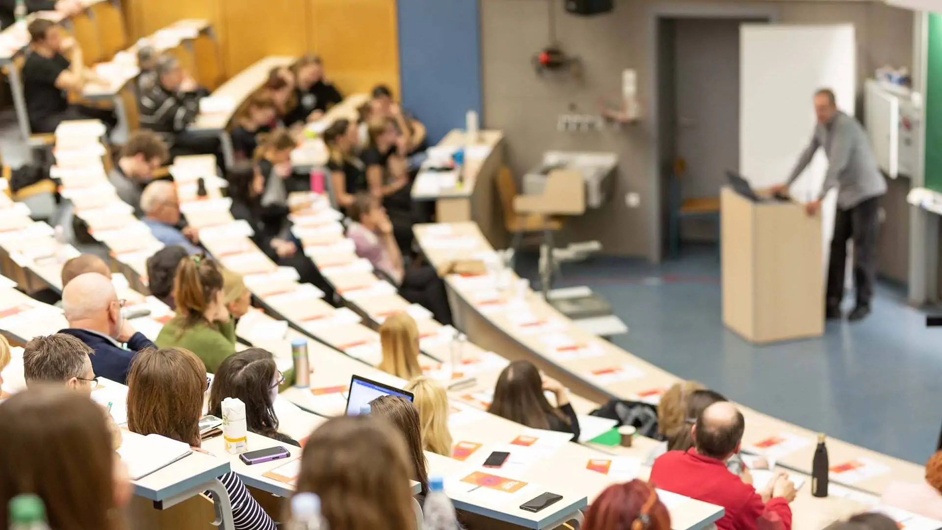 Aufnahme einer Vorlesung an einer Hochschule in einem Hörsaal mit aufsteigender Bestuhlung. EIn männlicher Dozent steht vorne am Rednerpult. Die Ränge sind mit Studierenden unterschiedlichen Geschlechts und Alters gefüllt.