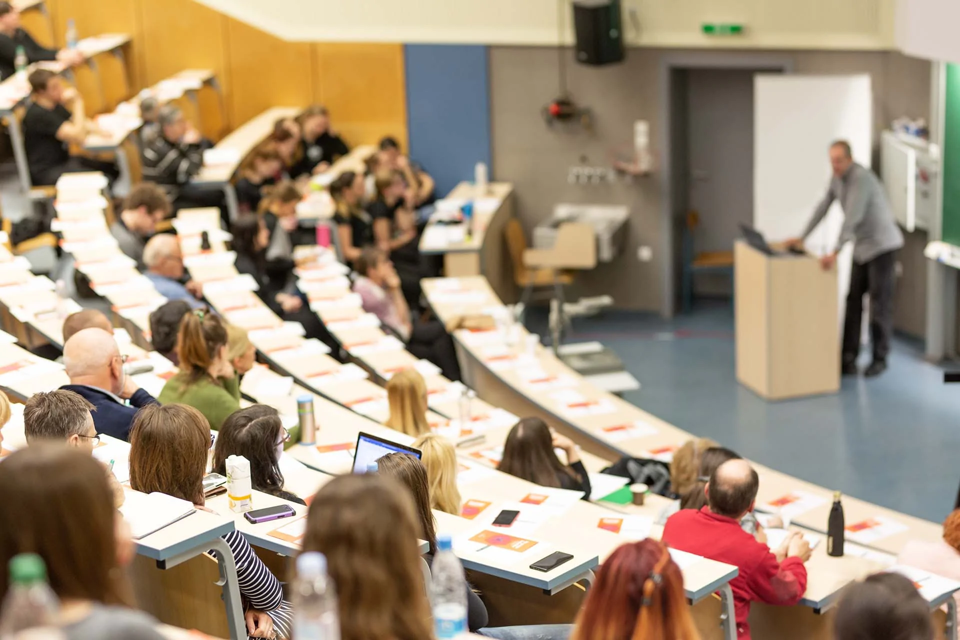 Aufnahme einer Vorlesung an einer Hochschule in einem Hörsaal mit aufsteigender Bestuhlung. EIn männlicher Dozent steht vorne am Rednerpult. Die Ränge sind mit Studierenden unterschiedlichen Geschlechts und Alters gefüllt.