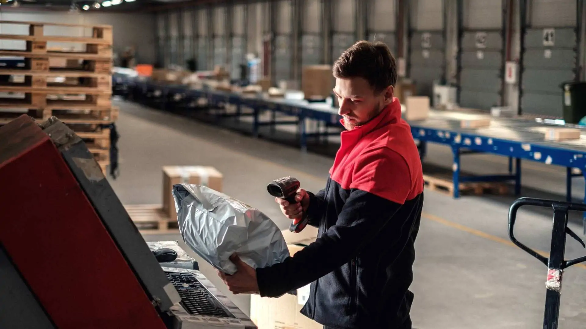 A warehouse worker in a large shipping hall scans a packaged parcel with a handheld scanner at a workstation with a computer. Conveyor belts, pallets, and other boxes can be seen in the background.