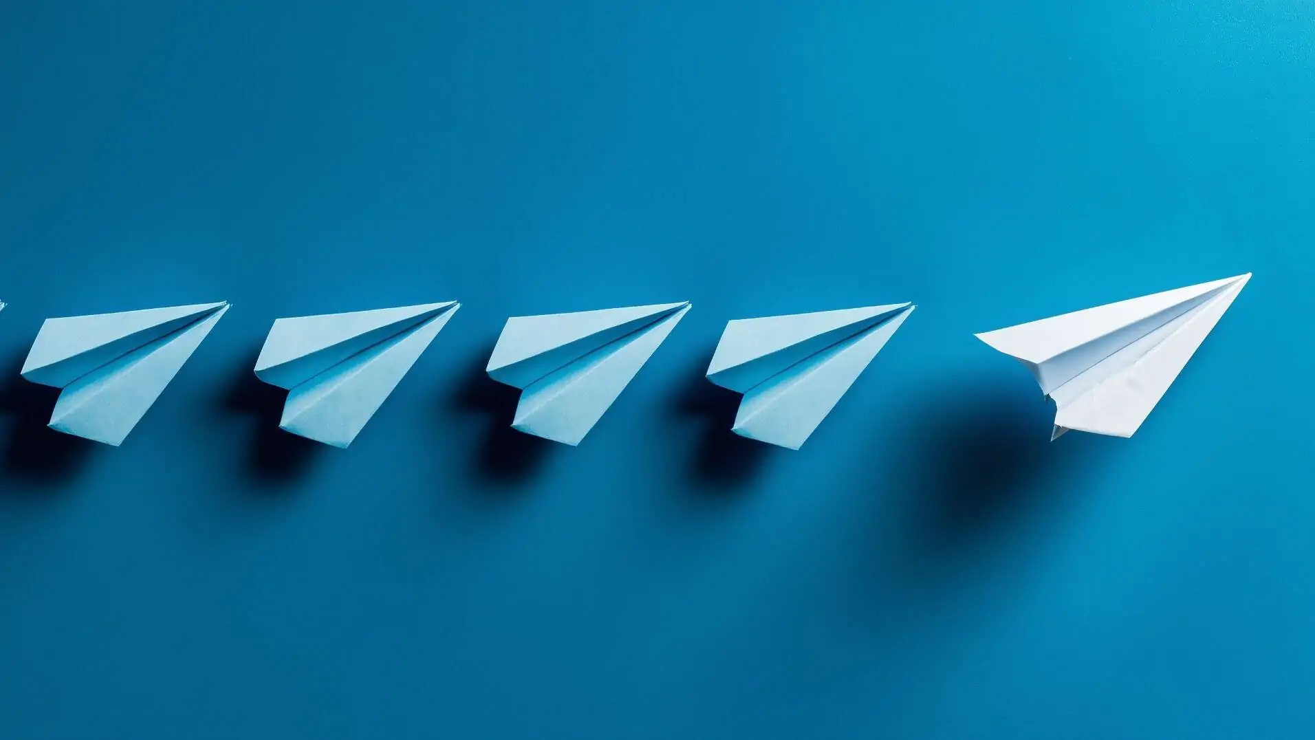 Vier blaue Papierflieger in einer horizontalen Reihe und rechts daneben leicht abgesetzt ein weisser Papierflieger
