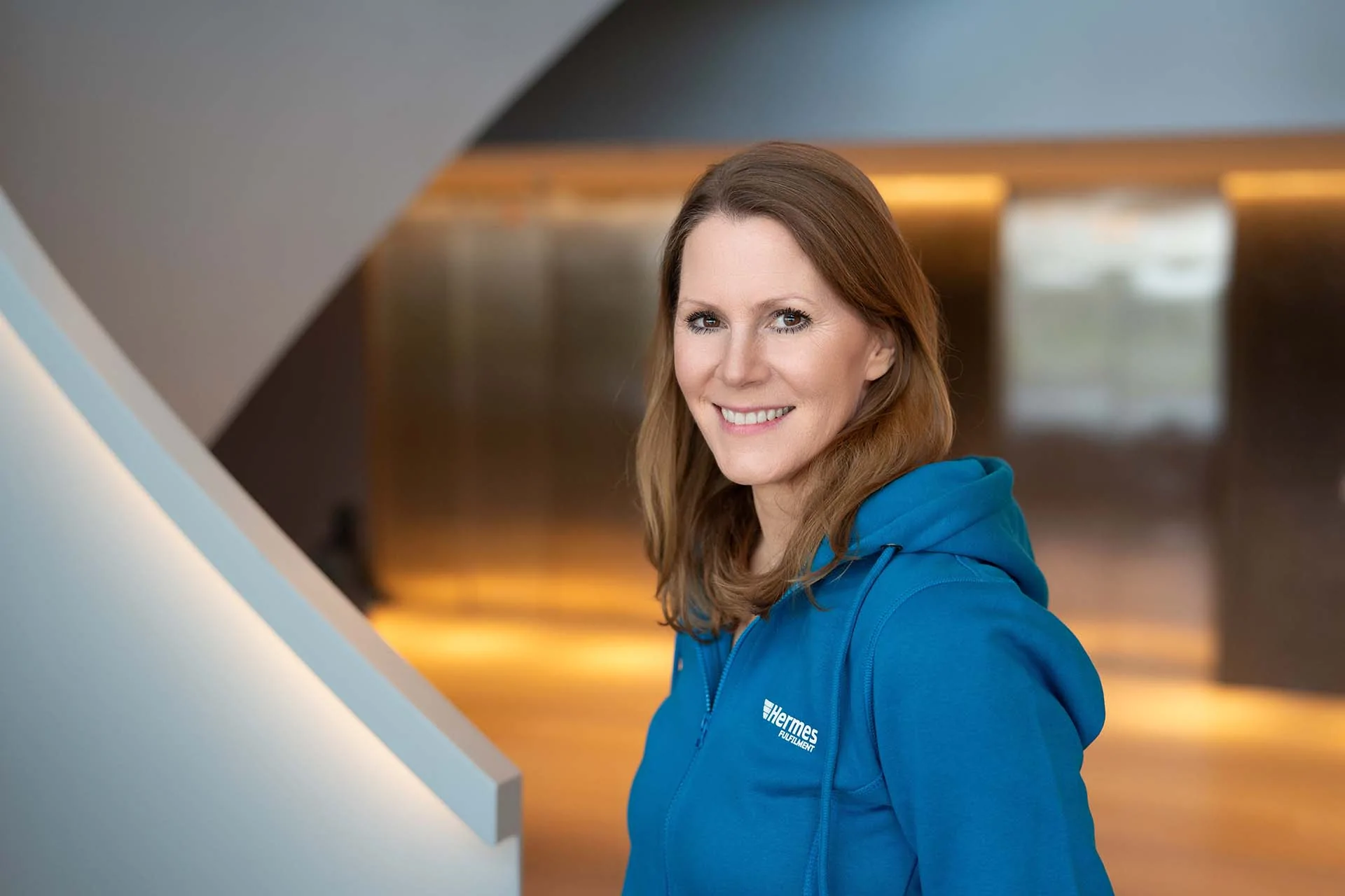 Corinna Falk&mdash;Head of Logistics Consulting and Planning for In-House Projects at Hermes Fulfilment. She is standing in front of a staircase in an office building and is wearing a blue hoodie with the brand name &ldquo;Hermes Fulfilment.&rdquo;