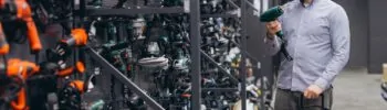 Ein Mann, der sich in einem Baumarkt ein neues Elektrowerkzeug aussucht. Werkzeuge in verschiedenen Farben sind Herstellerweise an einer Wand angebracht. Der Mann steht vor grünen Werkzeugen. Er hält ein grünes in den Händen und betrachtet es eingehend.
