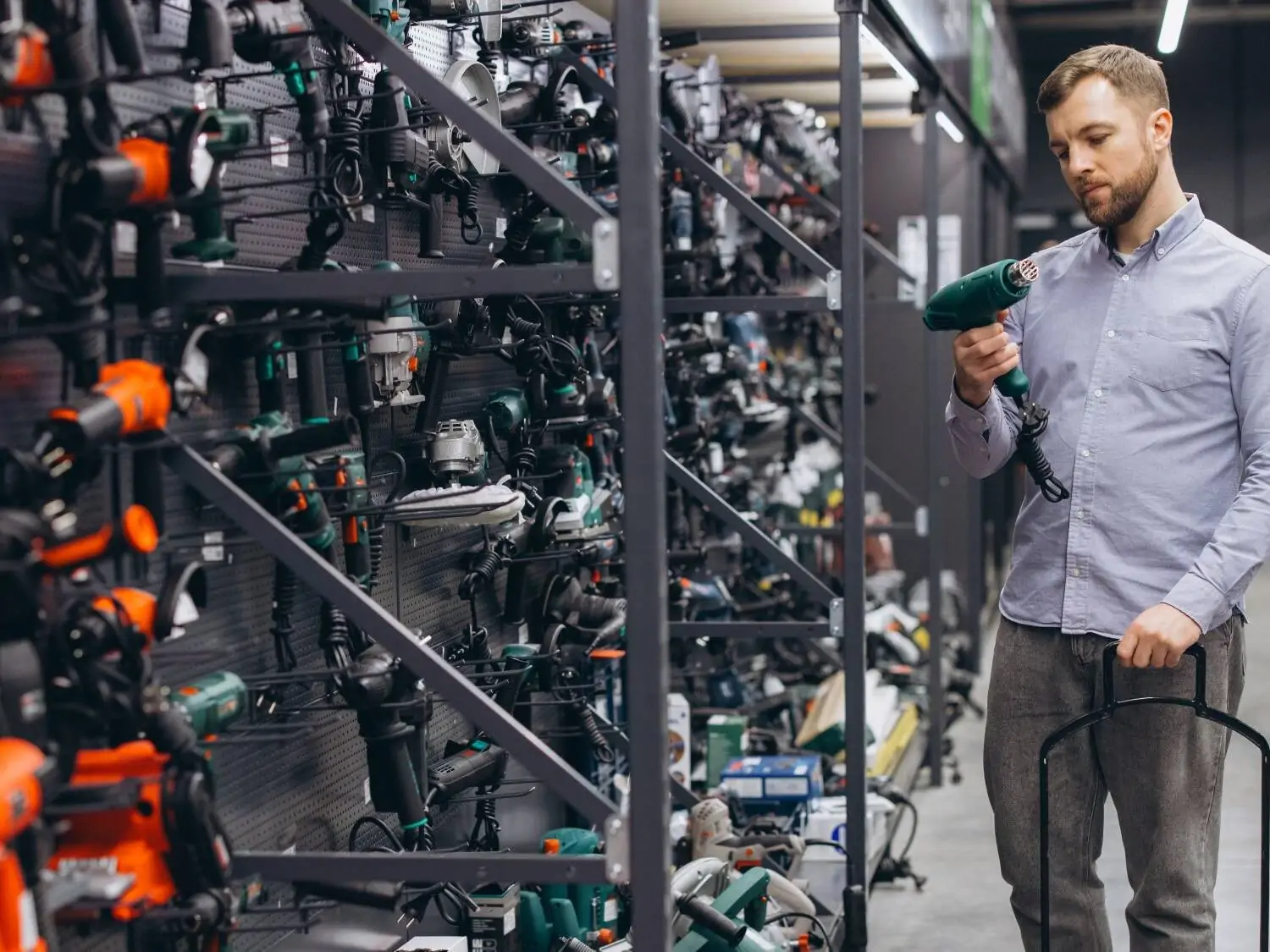 Ein Mann, der sich in einem Baumarkt ein neues Elektrowerkzeug aussucht. Werkzeuge in verschiedenen Farben sind Herstellerweise an einer Wand angebracht. Der Mann steht vor grünen Werkzeugen. Er hält ein grünes in den Händen und betrachtet es eingehend.