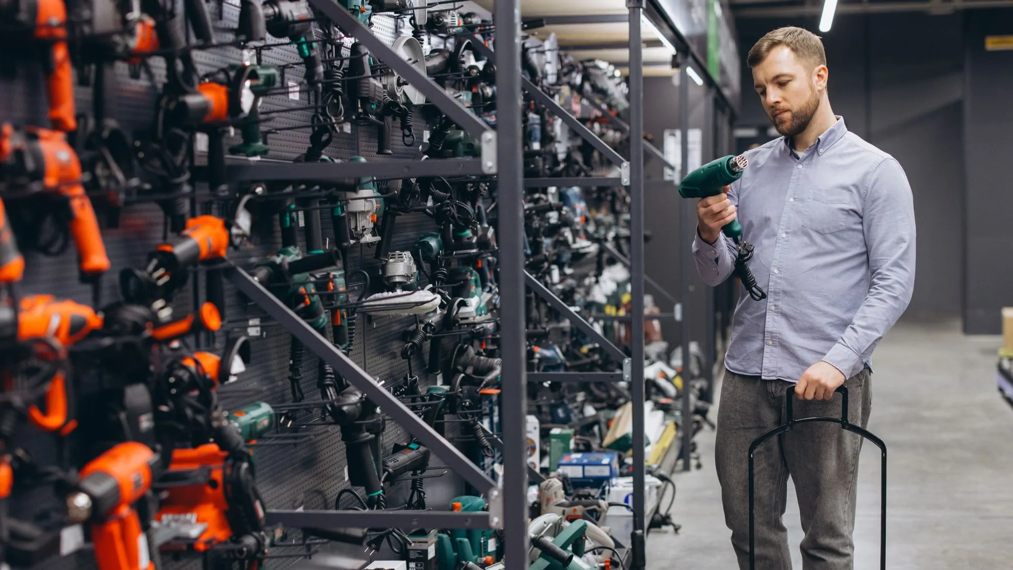 Ein Mann, der sich in einem Baumarkt ein neues Elektrowerkzeug aussucht. Werkzeuge in verschiedenen Farben sind Herstellerweise an einer Wand angebracht. Der Mann steht vor grünen Werkzeugen. Er hält ein grünes in den Händen und betrachtet es eingehend.